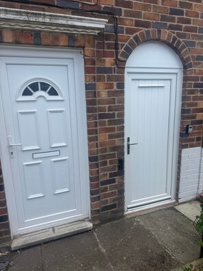 Two white doors with different designs, one with a semi-circular window and the other with a plain design, set against a brick wall with a white trim. - Knotty Ash - 3 Beds - Sleeps 6 - On-site Parking (Liverpool)