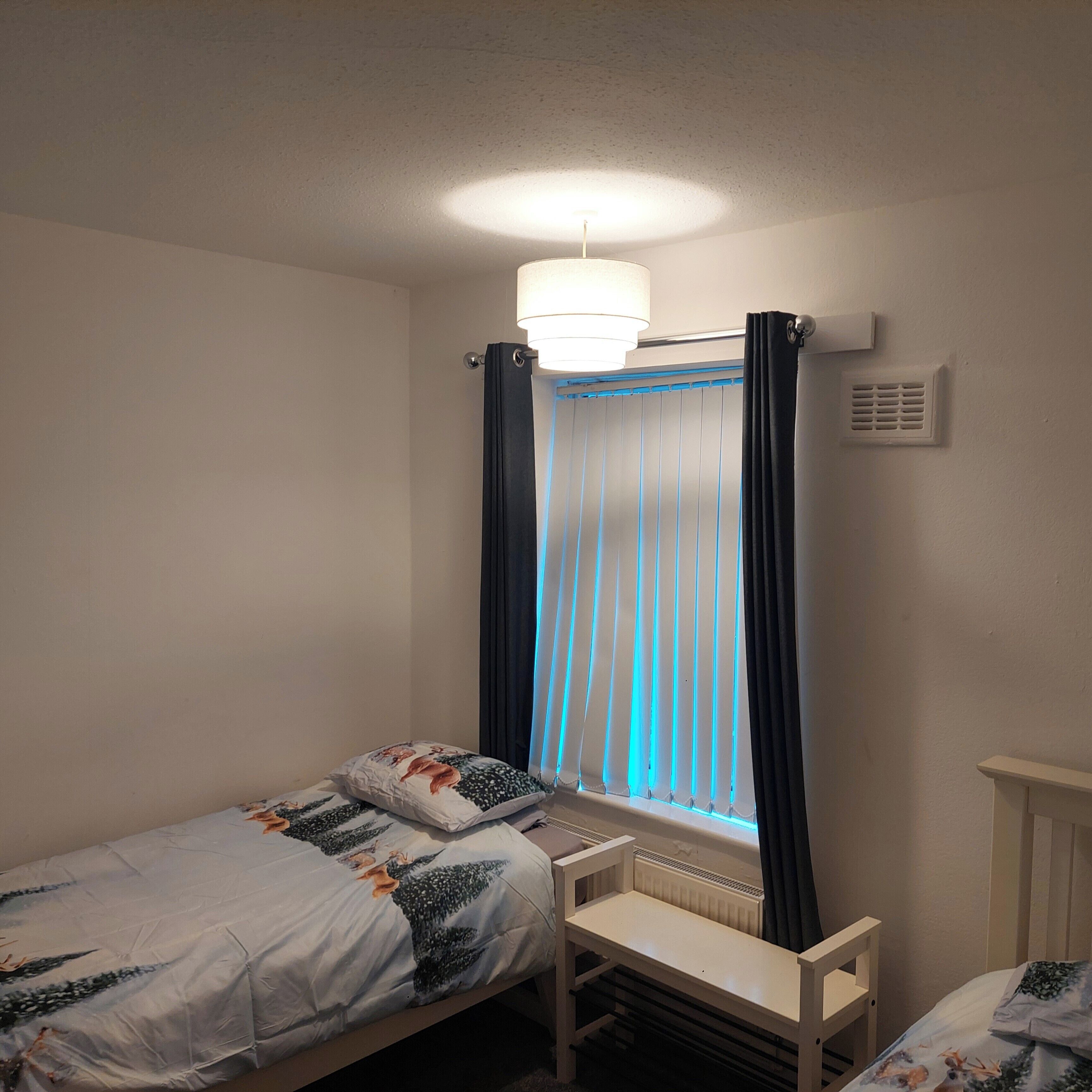A small, single-bed room with a window, a bedside table, and a radiator.