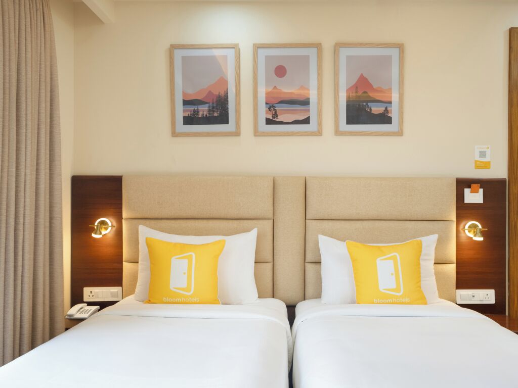 Habitación de hotel con dos camas, cada una con un colchón blanco y un almohadón amarillo con el logotipo del hotel. Encima de las camas hay tres cuadros enmarcados que representan paisajes.