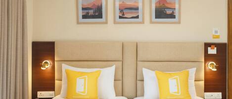 Habitación de hotel con dos camas, cada una con un colchón blanco y un almohadón amarillo con el logotipo del hotel. Encima de las camas hay tres cuadros enmarcados que representan paisajes.
