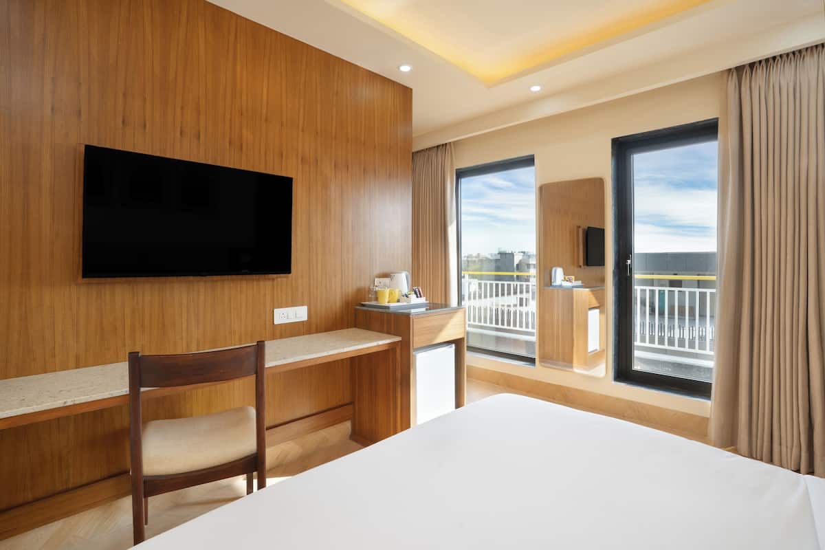 A hotel room with a bed, a desk, a chair, a television, a window with a view of the city, and a balcony.