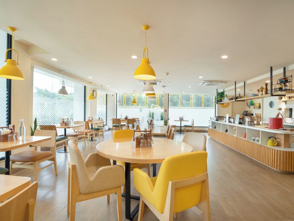 A modern cafe with round tables and yellow chairs, a wooden counter with various items, and large windows allowing natural light.