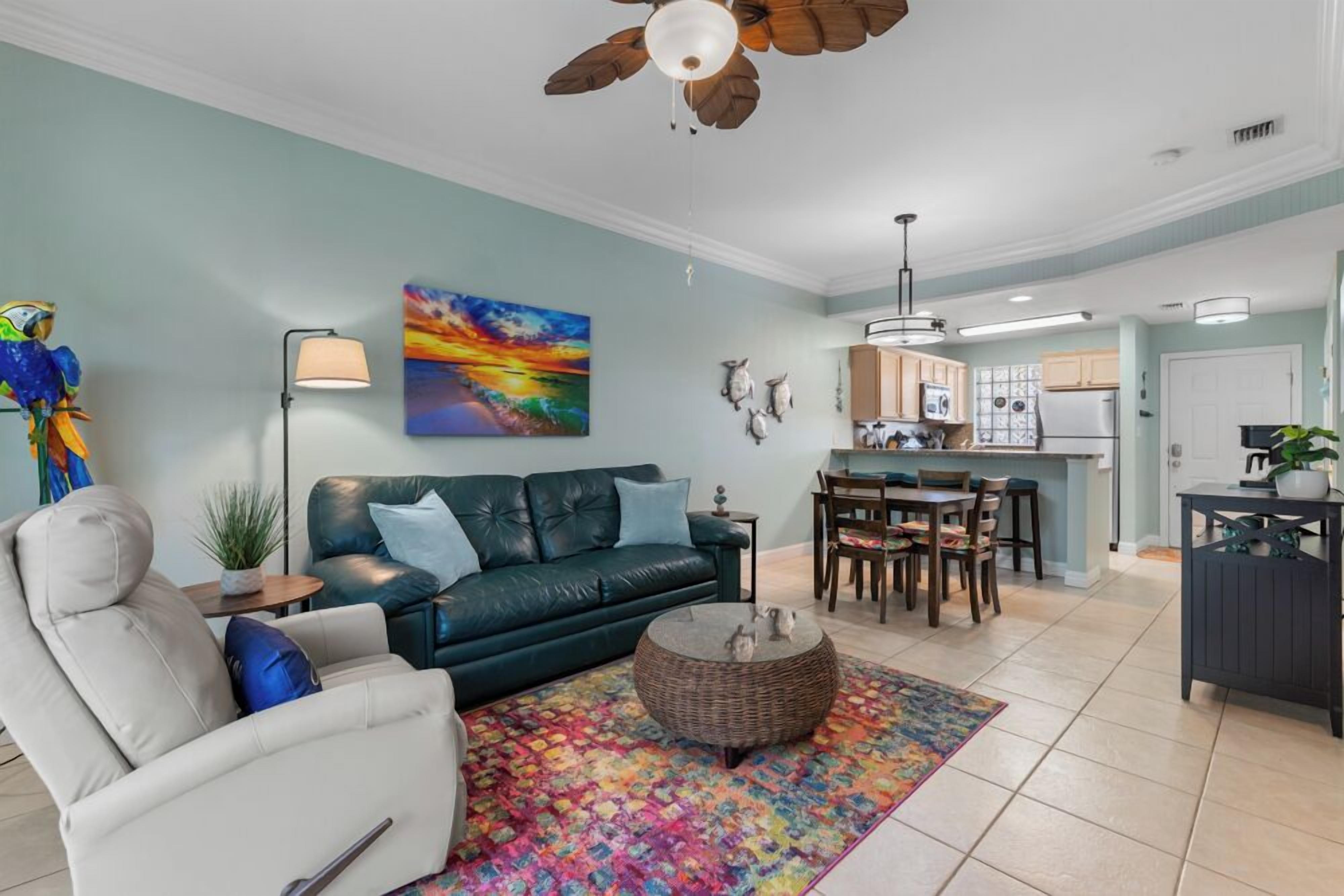 A living room with a sofa, armchair, coffee table, and a colorful rug. There is a dining area with a table and chairs.