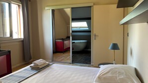 Une chambre d’hôtel avec un lit, un bureau, une chaise, une lampe et une salle de bain visible à travers une porte ouverte.