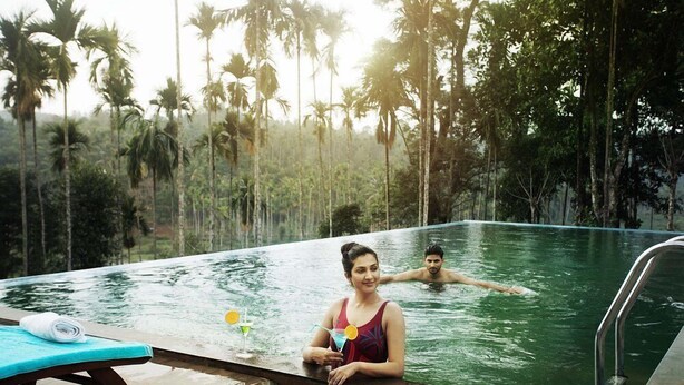 A swimming pool with two people in it, surrounded by tall palm trees. - The Wave Wayanad Resort (Sulthan Bathery)