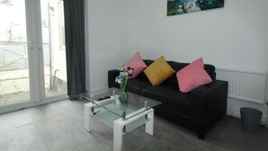 A modern living room with a glass coffee table, a black sofa with colorful cushions, a painting on the wall, and a view through a sliding glass door. - Sefton House by Smart Corporate Stays (Liverpool)