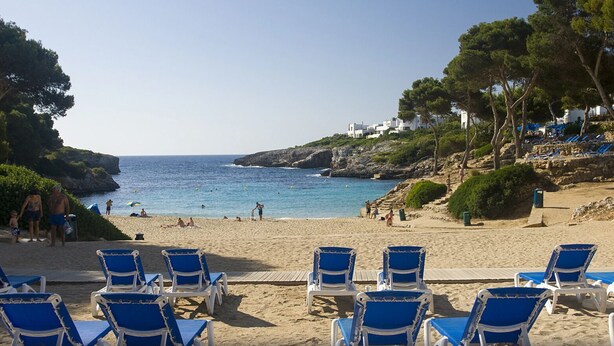 Una playa con sillas azules para tomar el sol, un litoral rocoso y edificios blancos al fondo.