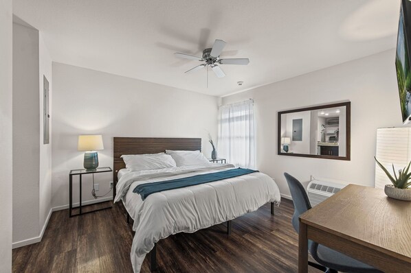 A bedroom with a bed, bedside table with a lamp, a ceiling fan, a dining table with a chair, and a mirror on the wall. - Overland Park Apartment King Bed, Workspace, & Gym (Overland Park)