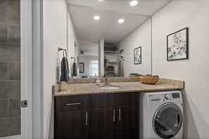 A modern bathroom with a built-in washing machine, dark wooden cabinets, a granite countertop, and a large mirror. - Overland Park Apartment King Bed, Workspace, & Gym (Overland Park)