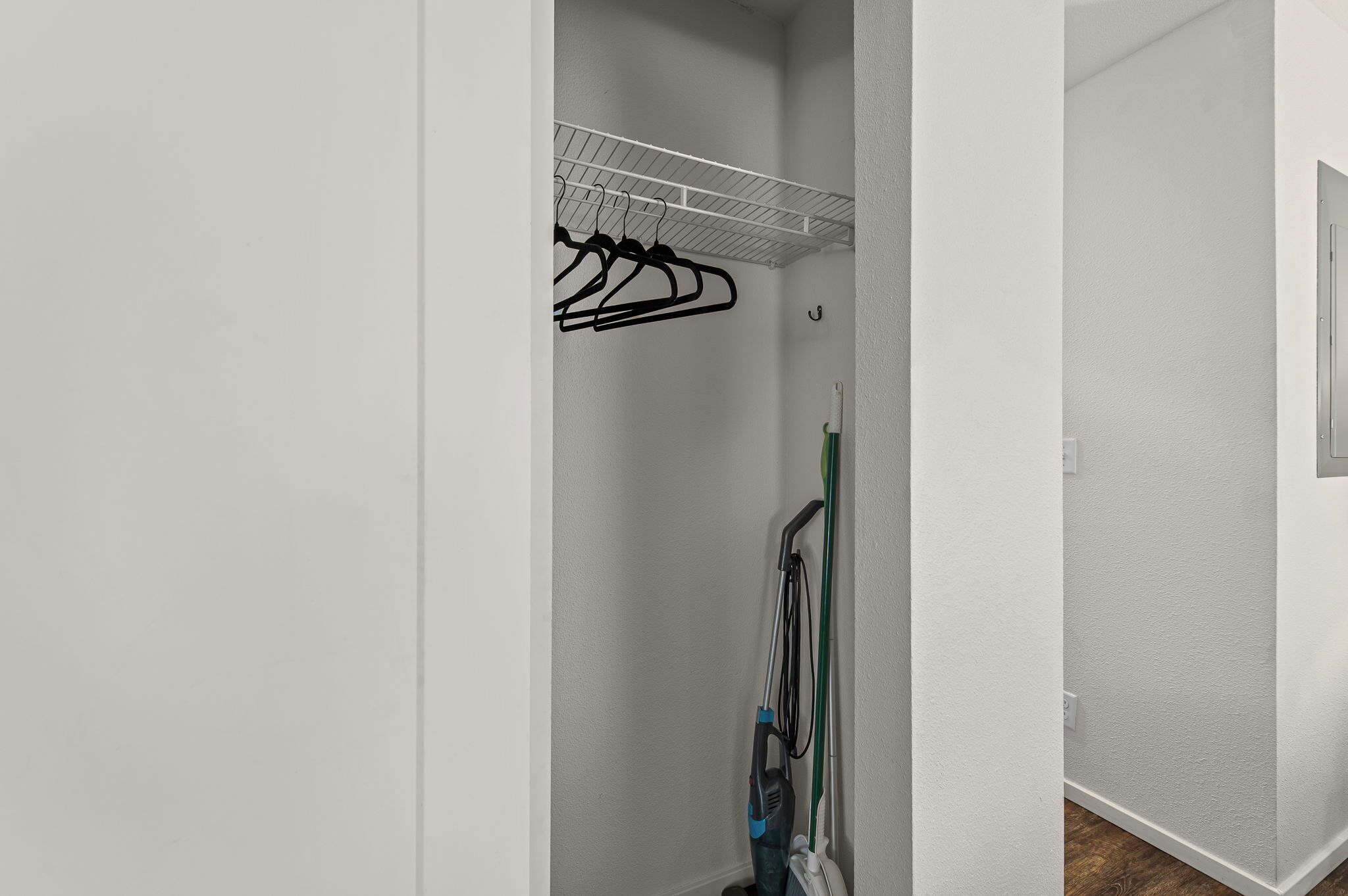 A narrow hallway with a white wall, a metal shelf with black hangers, and a green vacuum cleaner leaning against the wall.