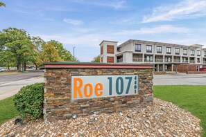 A sign for Roe 107 with a stone and metal design, located in front of a modern building with multiple windows and balconies. - Overland Park Apartment King Bed, Workspace, & Gym (Overland Park)