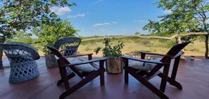 Une terrasse en bois, agrémentée de chaises et d’une table en osier, qui offre une vue sur un paysage verdoyant, avec des arbres et un ciel dégagé.