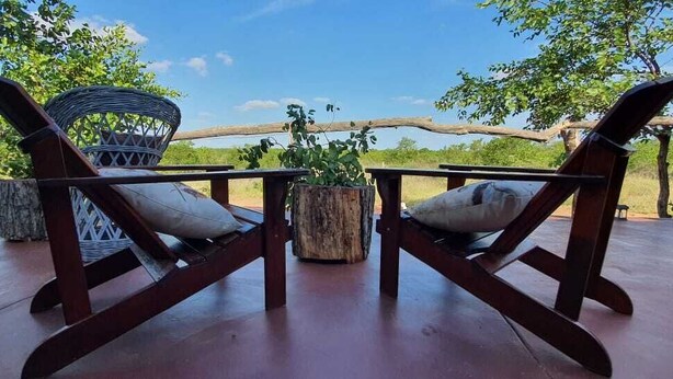Two wooden chairs with cushions, a woven basket, and a wooden stump table, all on a wooden deck with a view of trees and a clear sky. - Shanatsi Safari Lodge (Phalaborwa)