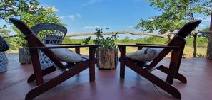 Deux chaises en bois avec des coussins, un panier en osier et une petite table en bois, le tout sur une terrasse en bois offrant une vue sur les arbres et un ciel dégagé.