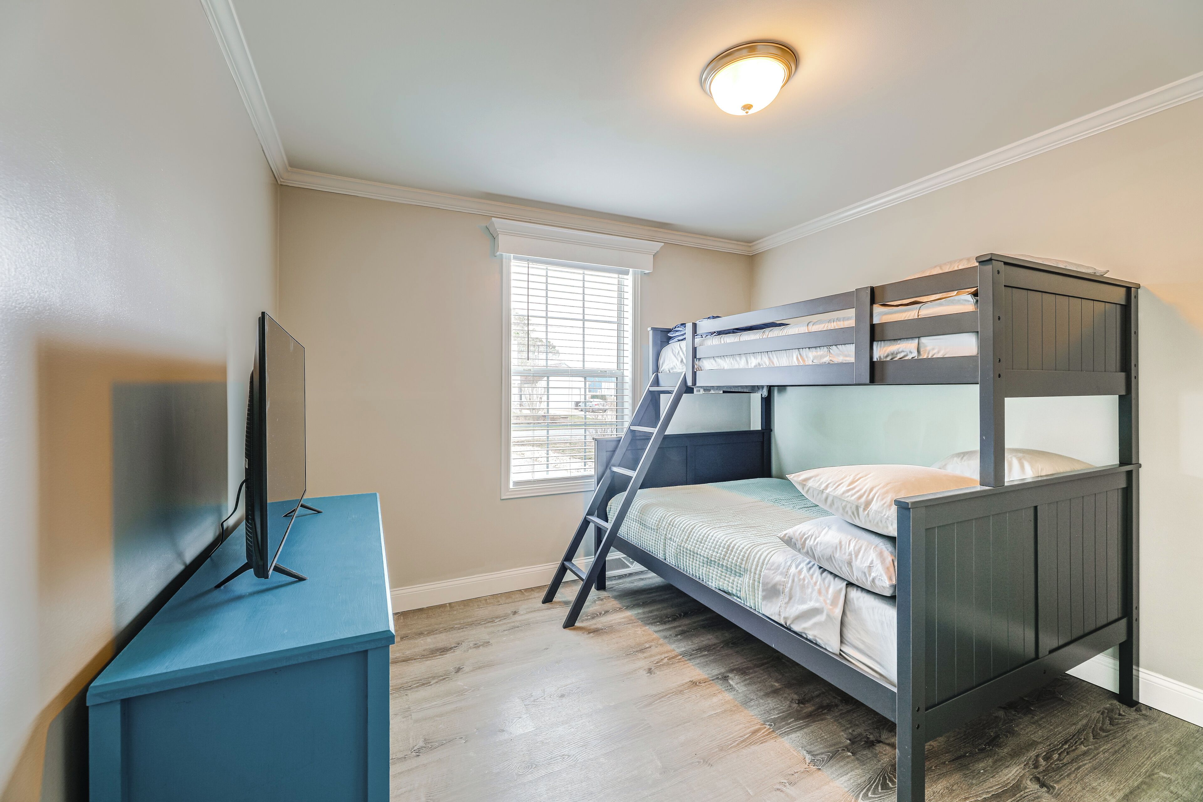 A bedroom with a bunk bed, a TV on a stand, a window with blinds, and a wooden floor.