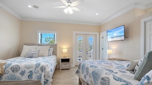 A bedroom with two beds, a ceiling fan, a TV, and a view of the outdoors through a door.