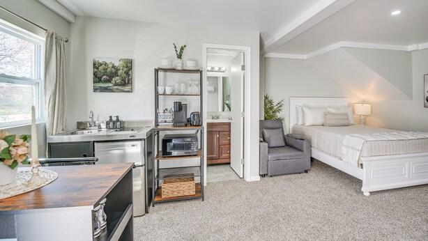 A modern bedroom with a bed, bedside table, and a grey armchair. A kitchen with a sink and cabinets is visible in the background. - 11 Mi to Virginia Beach Boardwalk: Studio Retreat! (Virginia Beach)