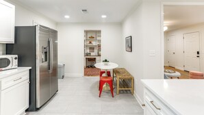 A modern kitchen with white cabinetry, a stainless steel refrigerator, a microwave, and a dining area with a round table and red stool.