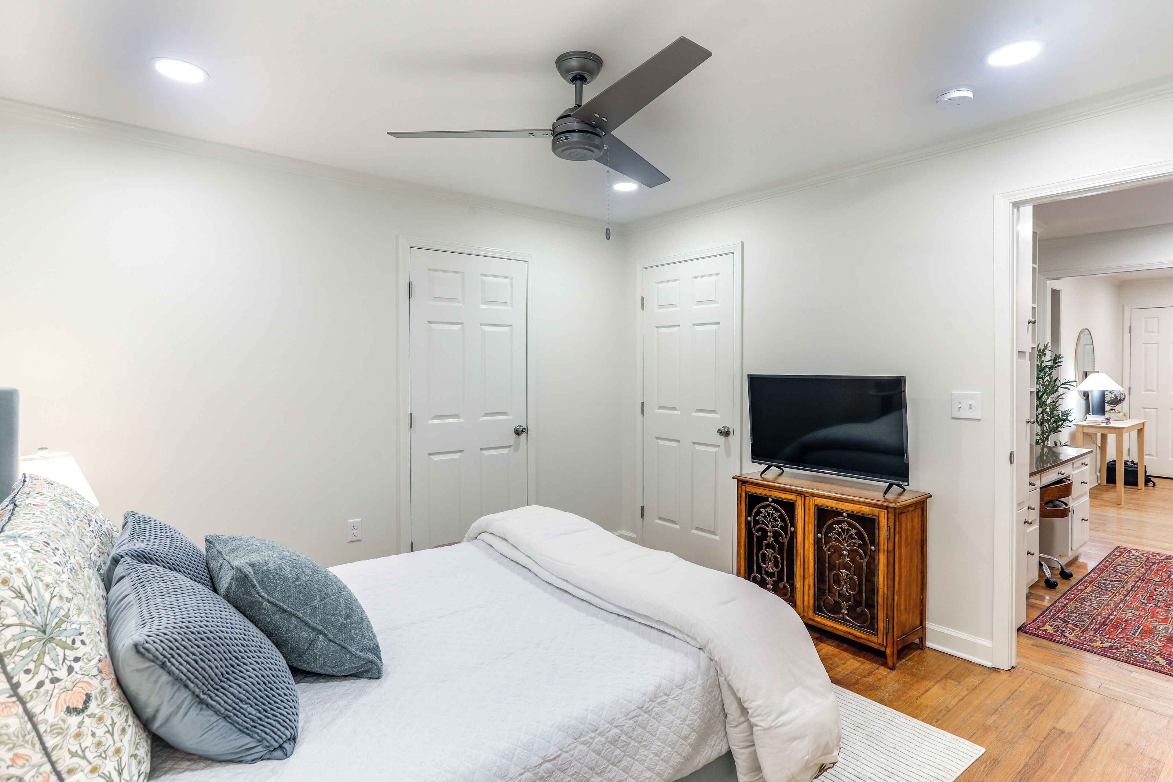 Une chambre à coucher avec un lit, un ventilateur de plafond, un téléviseur posé sur un meuble en bois et des portes blanches.