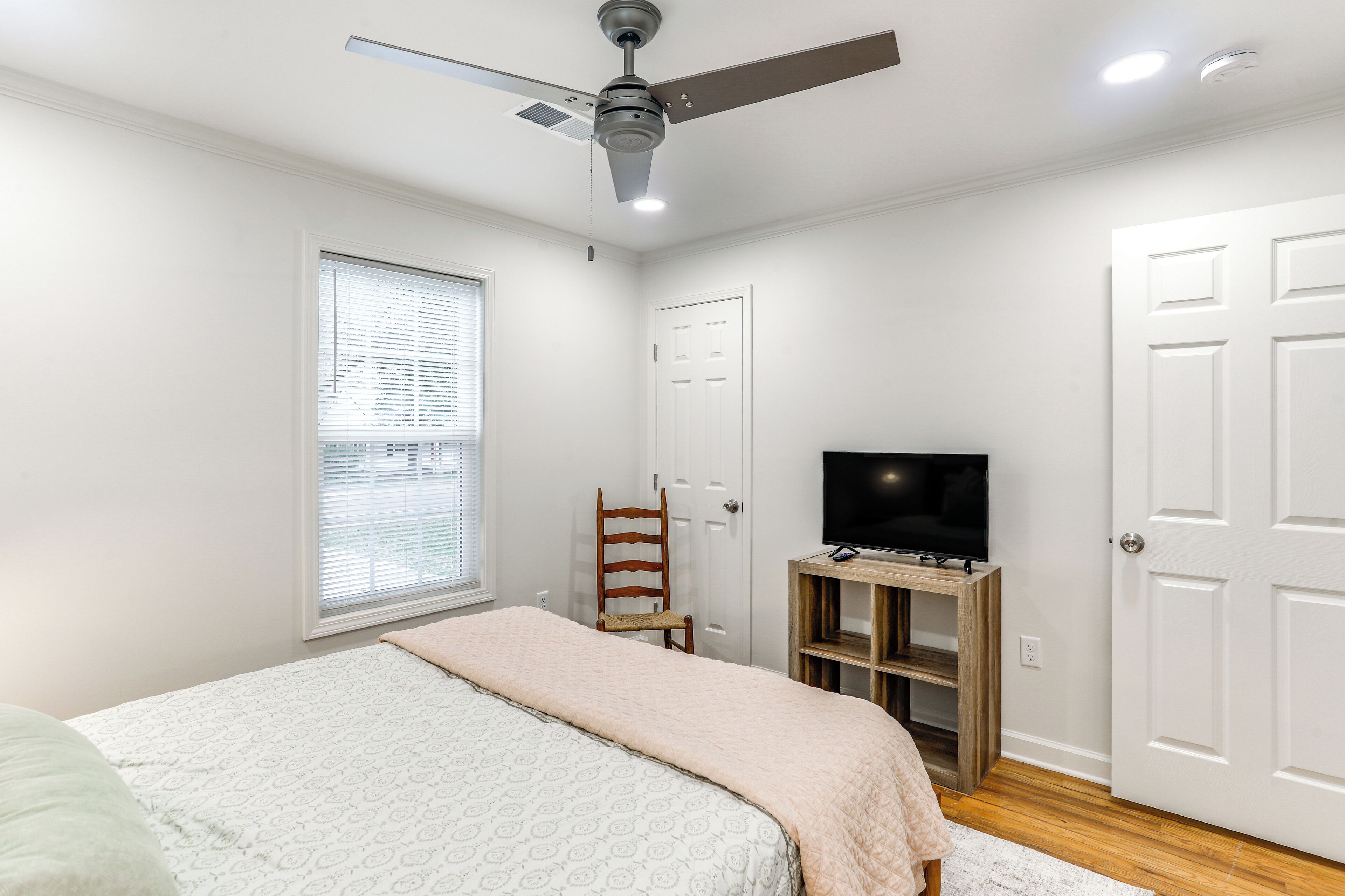 A bedroom with a bed, a TV on a wooden stand, a ceiling fan, a window with blinds, and a door.