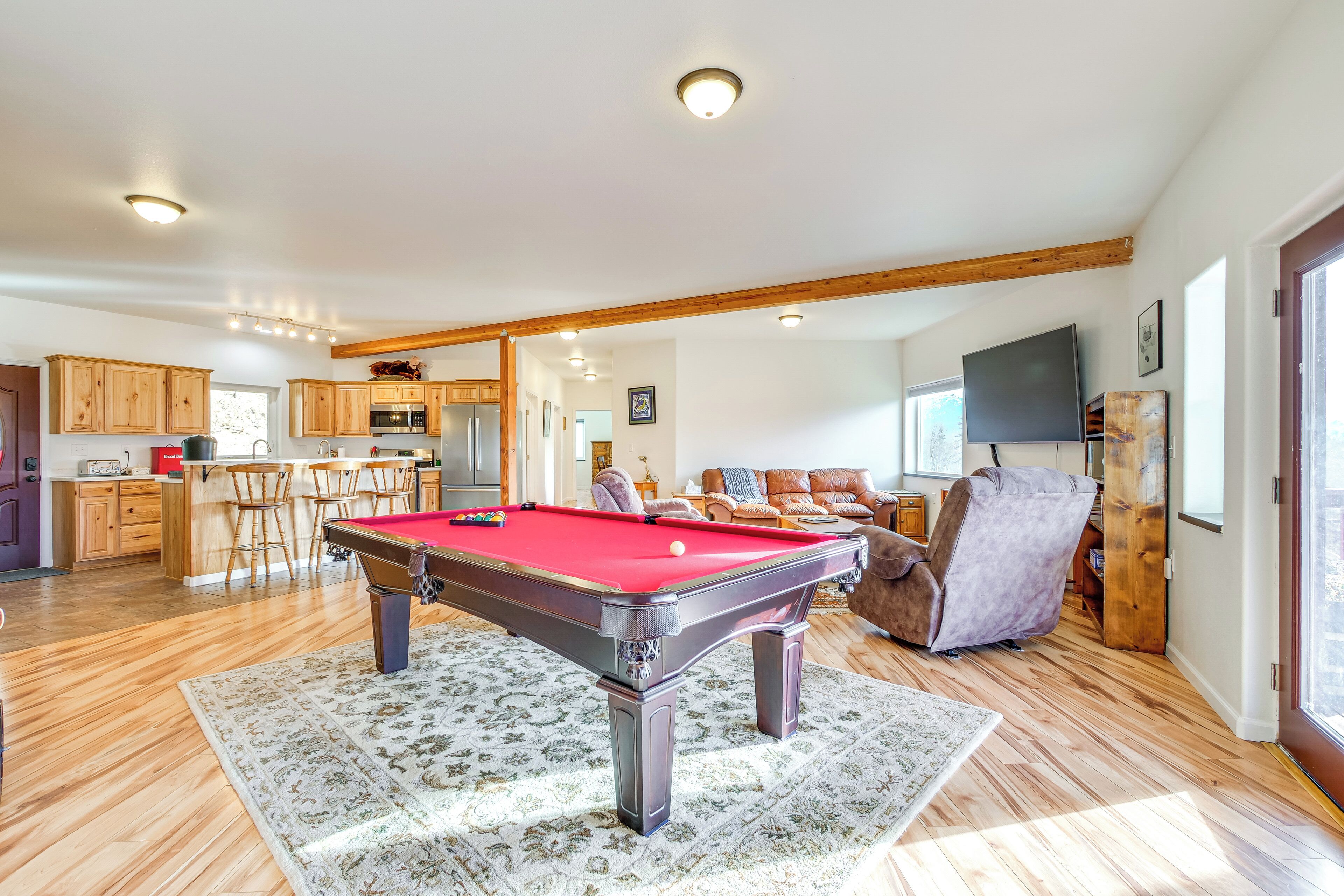 A pool table in a well-lit room with wooden flooring, a kitchen area, and a living room with a large TV.