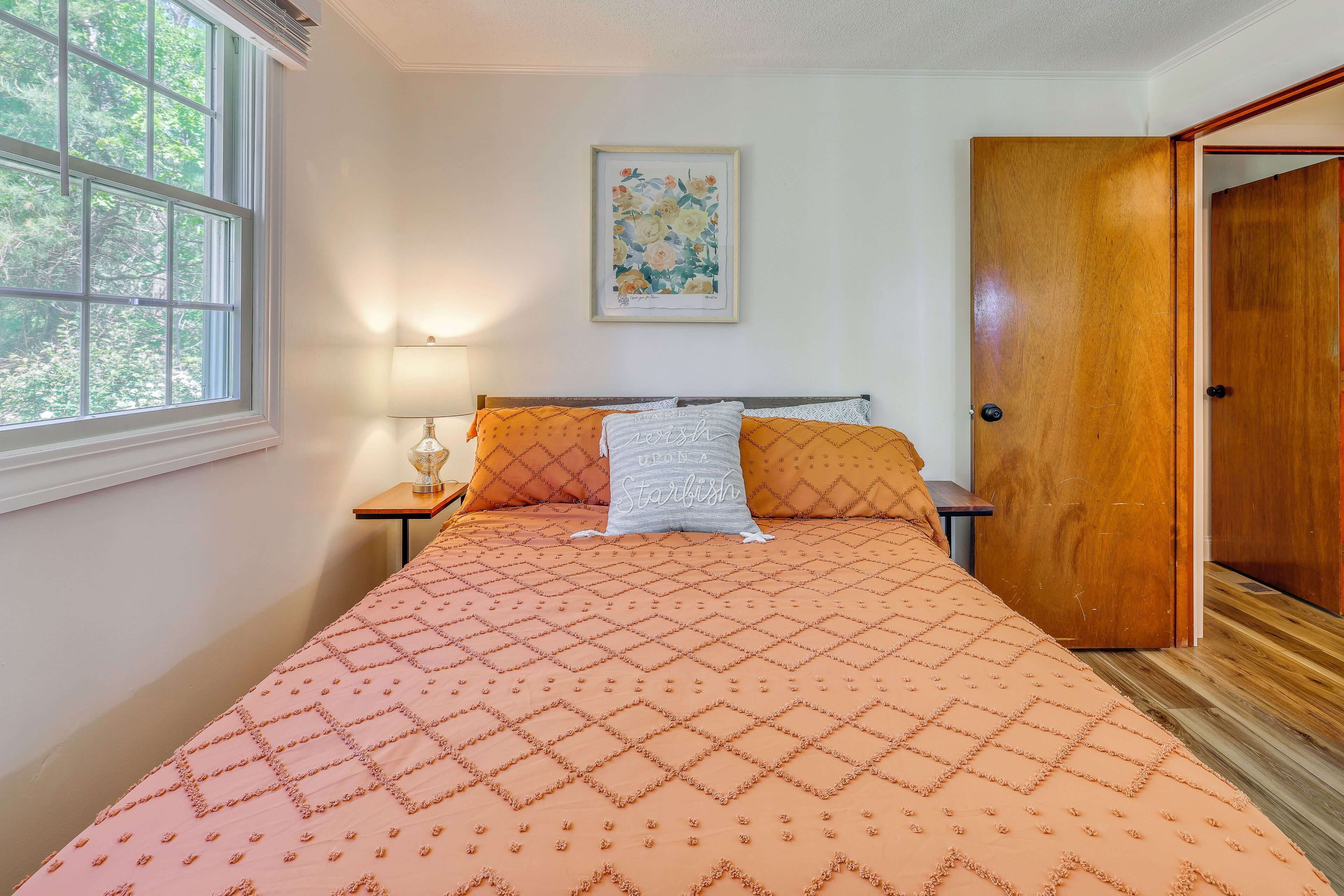 A bedroom with a bed, a nightstand, a lamp, a framed floral artwork, and a wooden door.