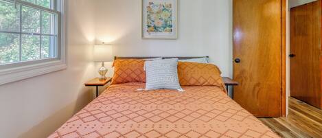 A bedroom with a bed, a nightstand, a lamp, a framed floral artwork, and a wooden door.