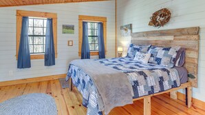 Une chambre à coucher avec un lit en bois, deux fenêtres avec des rideaux, une tête de lit en bois et un élément décoratif fixé au mur.