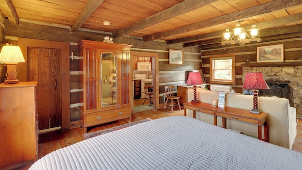 A log cabin bedroom with a wooden ceiling, a large bed, a wooden dresser, a cabinet, a chair, and a fireplace.