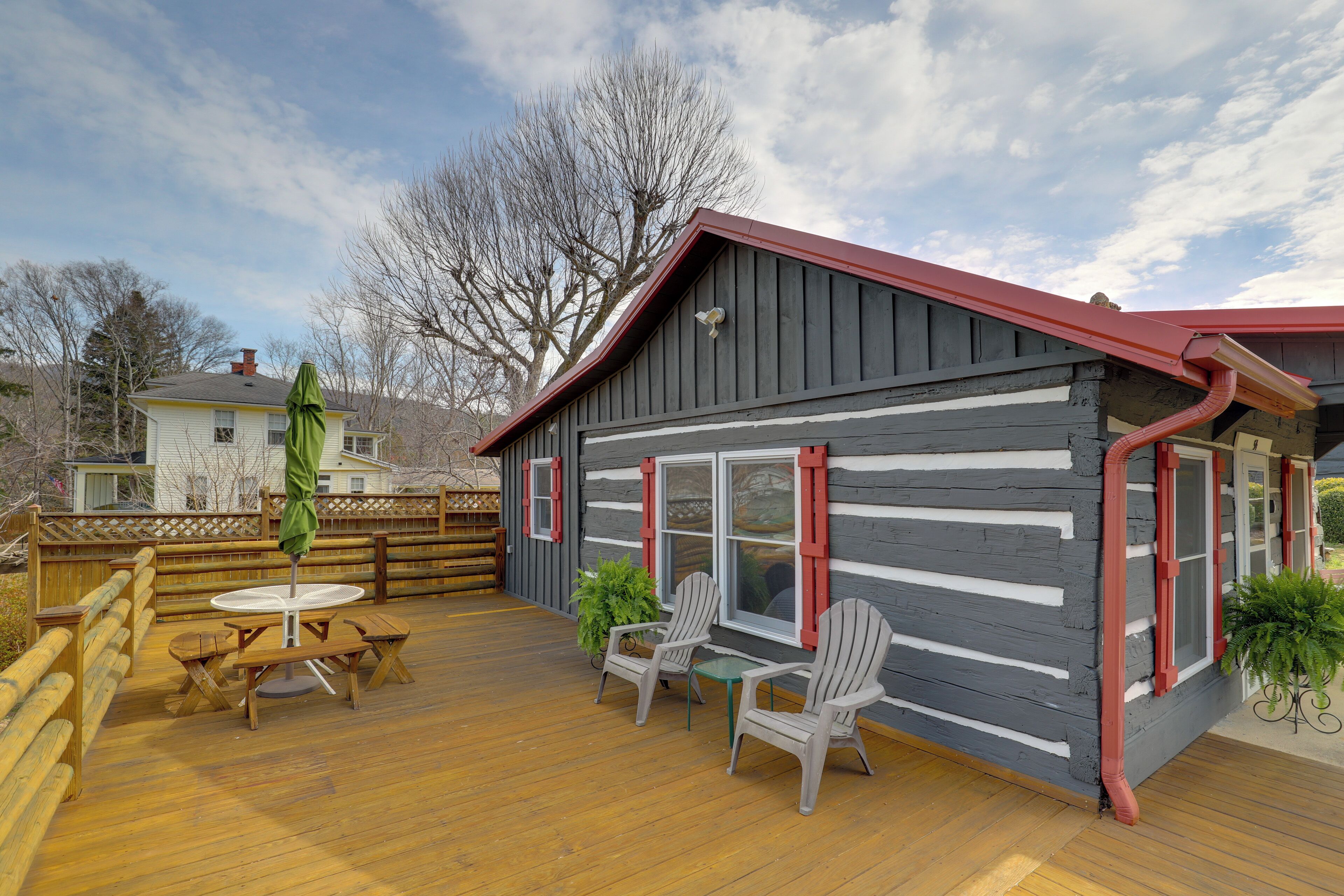 A wooden deck with a table and chairs, a small building with red trim, and a view of a house in the background.