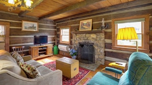 A log cabin interior with a stone fireplace, wooden walls, a television, and a cozy seating area.