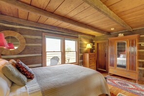 A log cabin bedroom with a large bed, wooden ceiling beams, a wooden wardrobe, and a view through a sliding glass door. - Scenic Hot Springs Retreat Near Appalachian Trail! (Hot Springs)