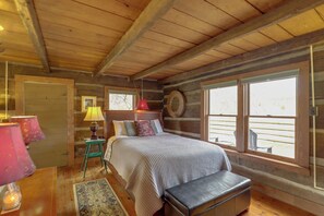 A cozy cabin-style bedroom with a wooden ceiling, a large bed with a headboard, a bench, a small table with a lamp, and a window with a view of the outside. - Scenic Hot Springs Retreat Near Appalachian Trail! (Hot Springs)