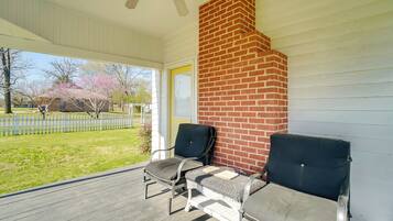 A covered porch with two chairs, a brick fireplace, and a view of a yard with trees and a house.