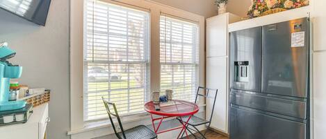 A compact kitchen with a stainless steel refrigerator, a red metal table with chairs, and a window with blinds.