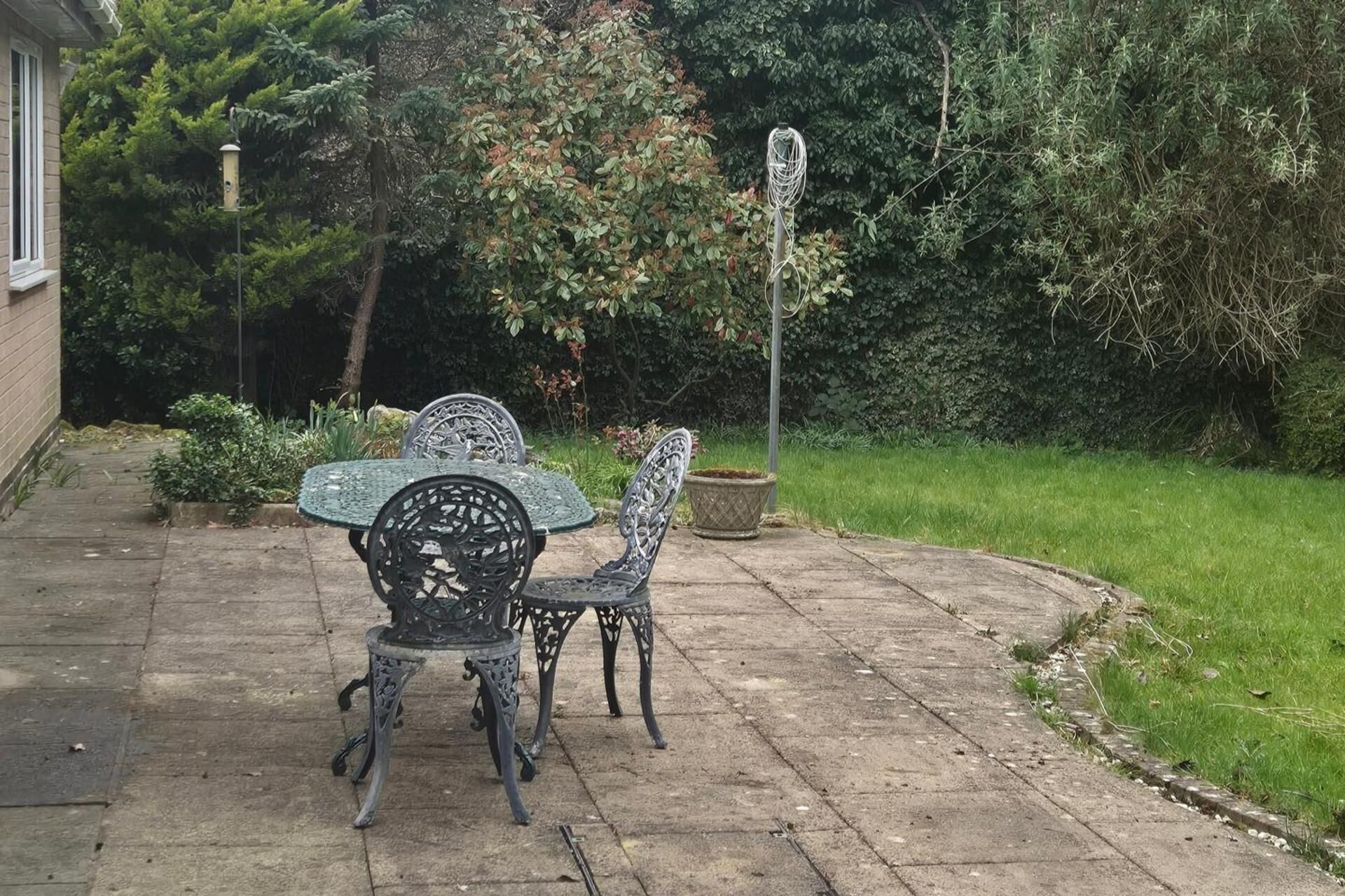 A patio with two ornate metal chairs and a table, surrounded by a garden with a variety of plants and a tall, thin pole.