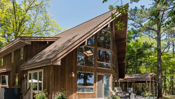 Una cabaña moderna de madera con ventanas amplias, una terraza con mobiliario exterior y un bosque de fondo.