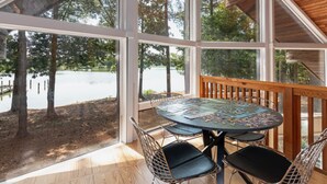 Un espacio al aire libre cubierto con una terraza de madera, una mesa redonda con sillas y amplios ventanales con vistas a un lago y árboles.
