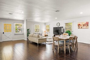 A spacious living room with a sofa, dining table, and TV. There are windows with blinds and a door with a decorative glass panel.