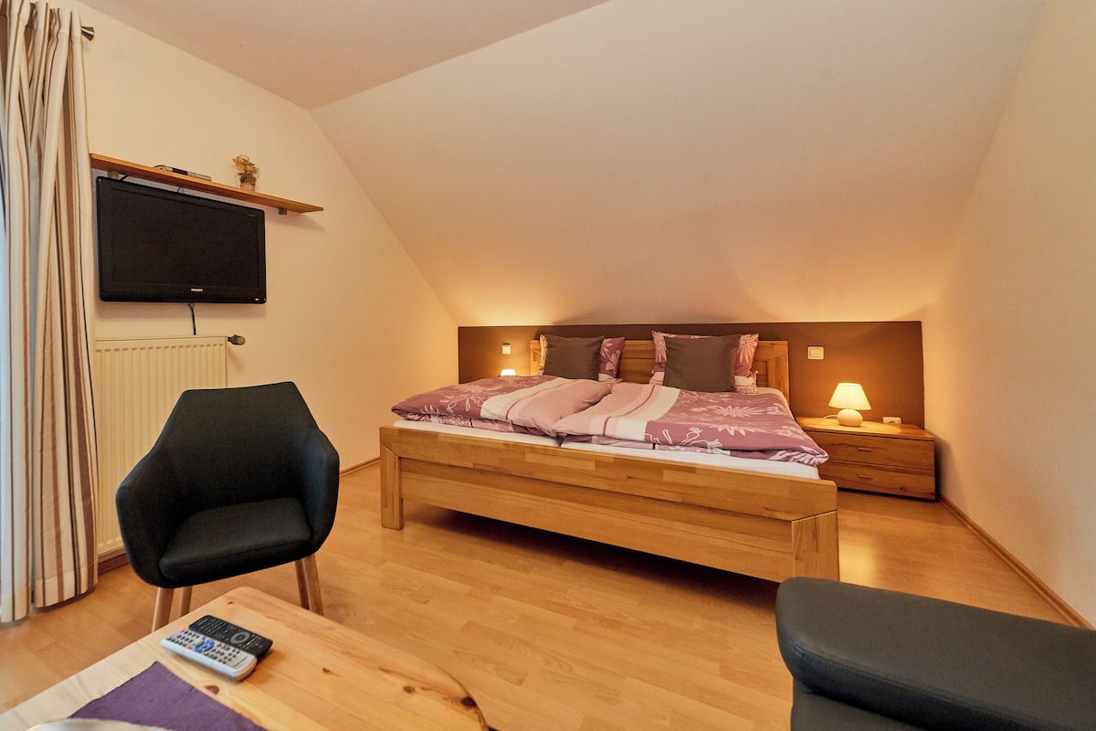 A bedroom with a bed, a chair, a nightstand, a lamp, and a TV mounted on the wall.