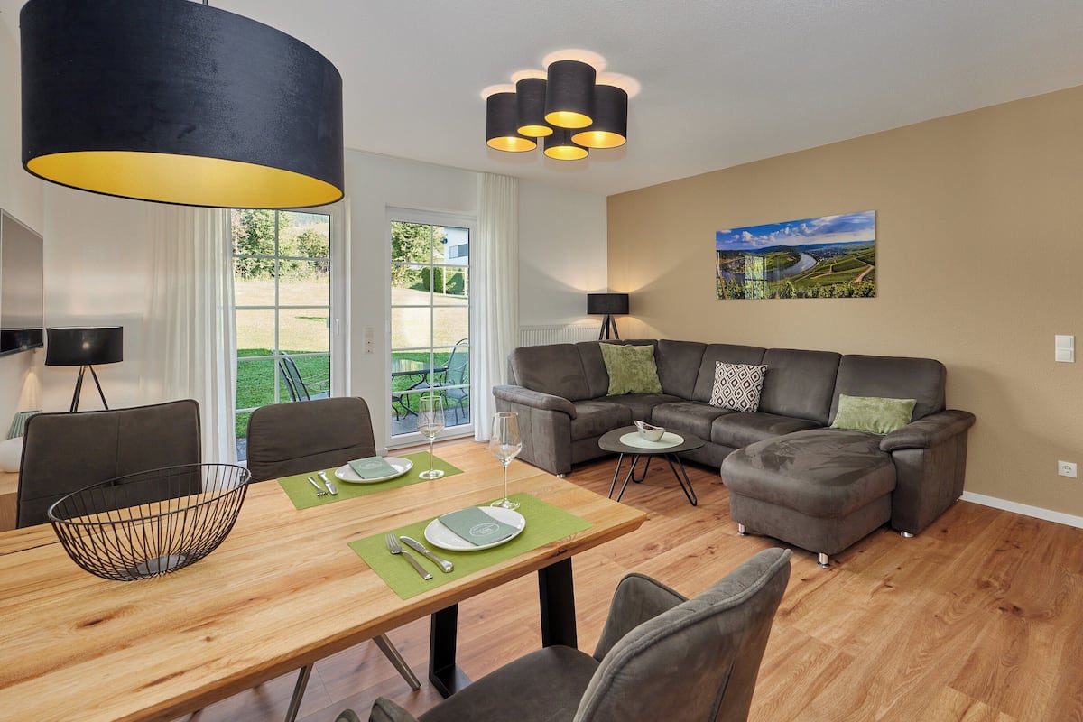 A modern living room with a wooden dining table, grey sofa, and a black hanging lamp.