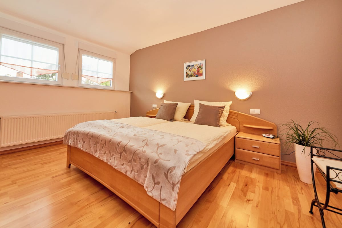 A bedroom with a wooden bed, two bedside tables, a nightstand, a potted plant, and a window with blinds.