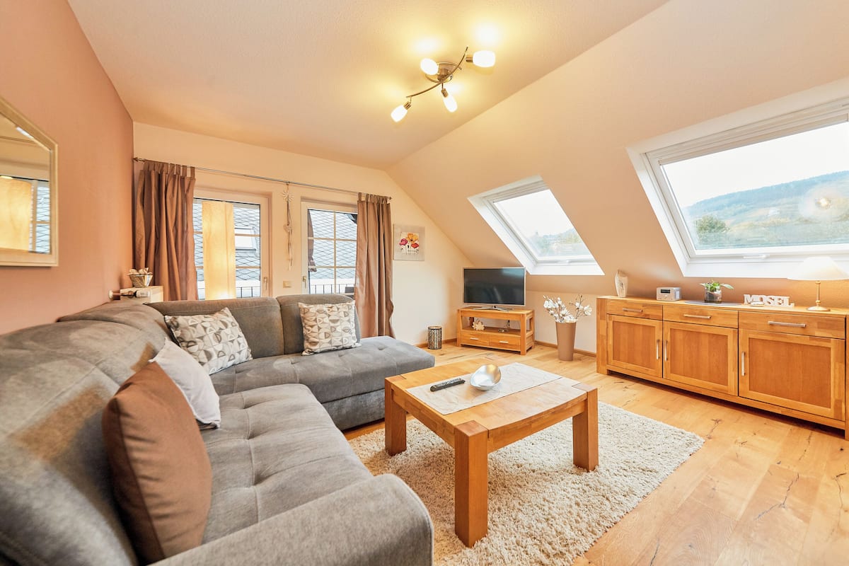 A modern living room with a sectional sofa, a wooden coffee table, and a TV on a wooden cabinet. There are two windows providing natural light and a view of the outdoors.