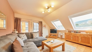 A modern living room with a sectional sofa, a wooden coffee table, and a TV on a wooden cabinet. There are two windows providing natural light and a view of the outdoors.