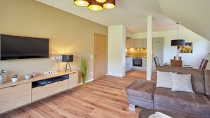 A modern living room with a flat-screen TV, wooden flooring, a brown leather sofa, and a dining area in the background.