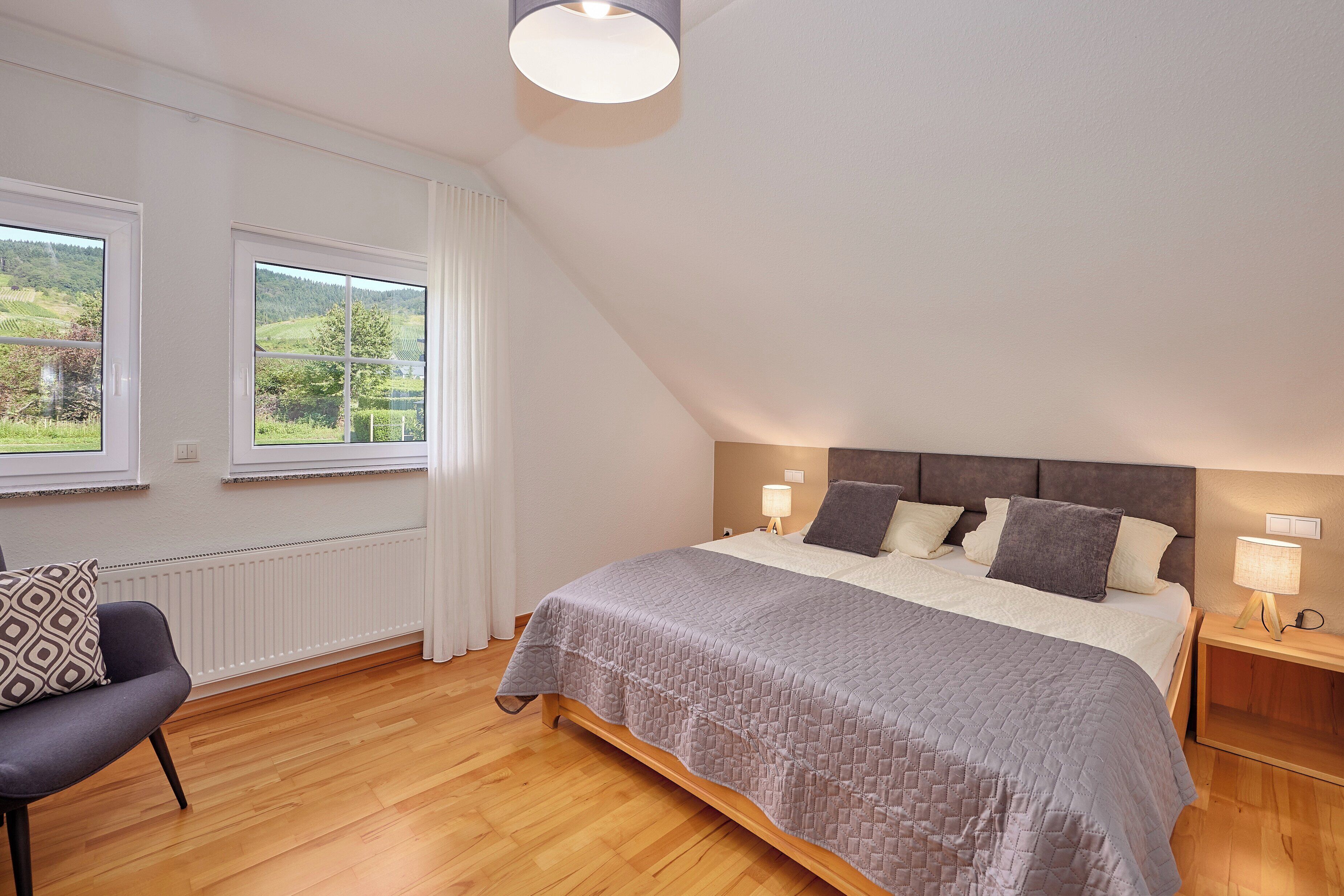 A bedroom with a large bed, two bedside lamps, a chair, and a view of greenery through the windows.