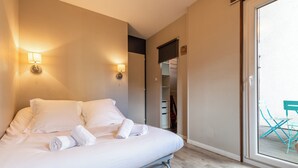 A neatly made bed with white linens and towels, a wall-mounted light fixture, and a sliding glass door leading to an outdoor area with a chair.