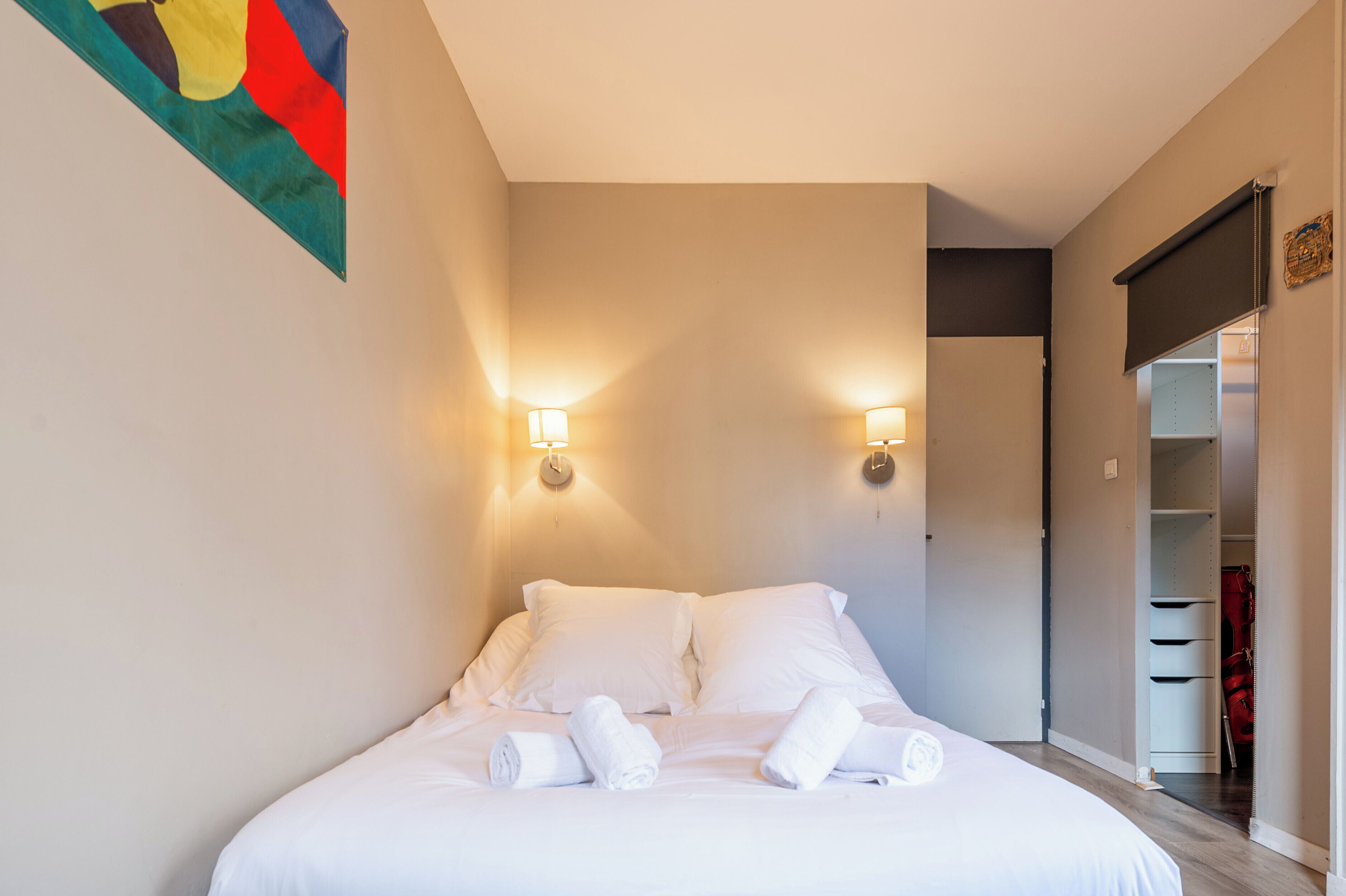 A neatly made bed with white linens and rolled towels, two wall-mounted lamps, and a closet with hanging clothes.