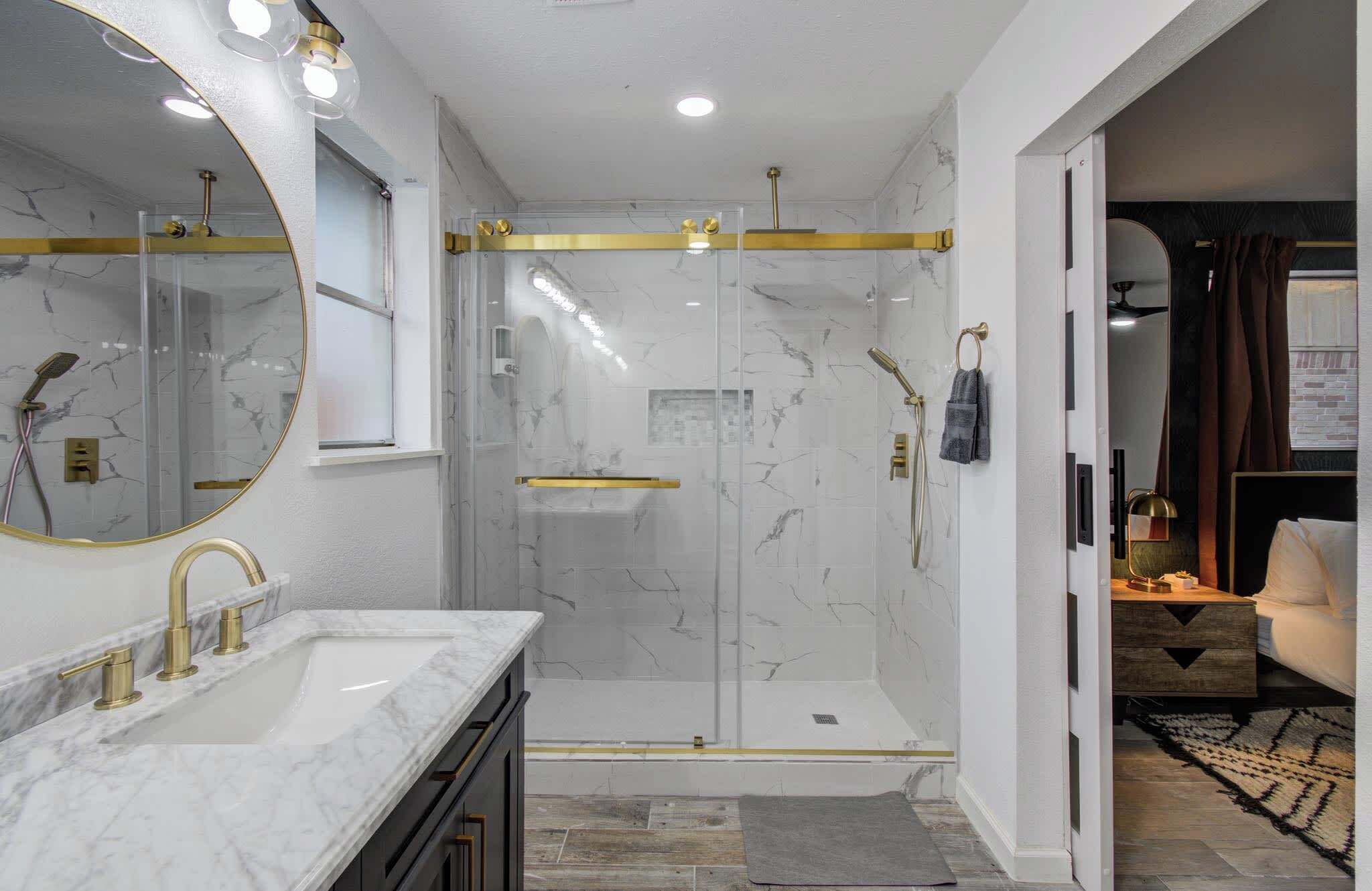 A modern bathroom with a marble vanity, a glass-enclosed shower, and a gold-framed mirror.