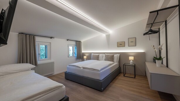 Quarto de hotel moderno com uma cama grande, duas mesas de cabeceira, uma mesinha lateral, um radiador e uma janela com cortinas.
